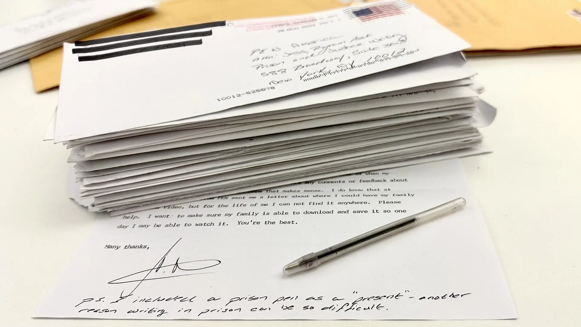 A stack of handwritten prison letters mail lies on a desk, with an uncapped pen resting on a typed letter. The letter mentions including a prison pen as a present and discusses the challenges of writing from prison.
