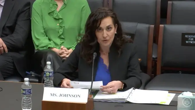 A woman with curly brown hair, wearing a blue top and black blazer, delivers PEN America testimony at a panel with a microphone. A nameplate reading Ms. Johnson and documents are in front of her, alongside two water bottles on the table.