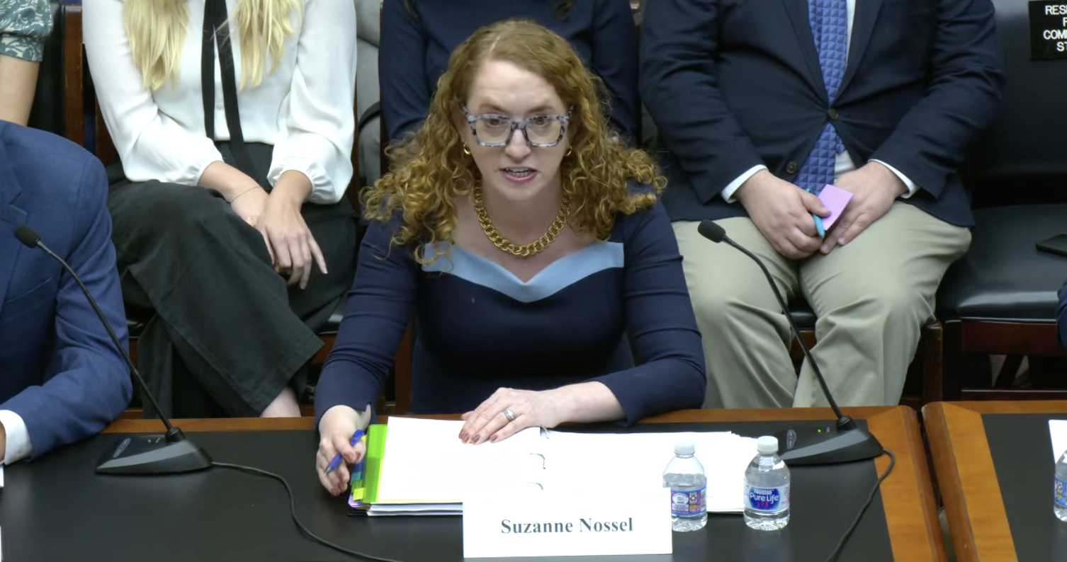 A woman with curly red hair, glasses, and a blue dress delivers PEN America testimony at a desk with an open binder, two water bottles, and a nameplate reading “Suzanne Nossel.” People are seated behind her.