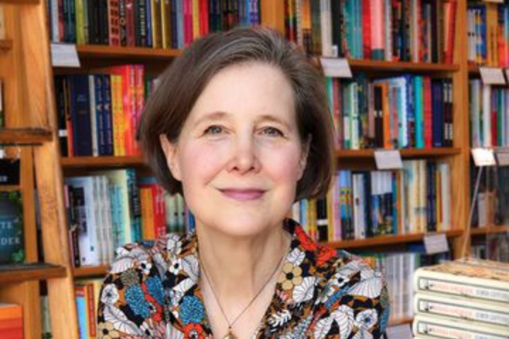 A woman with short brown hair and a floral shirt smiles softly while sitting in front of shelves filled with colorful books, perhaps browsing for an Ann Patchett novel that thoughtfully explores the impact of a school shooting.