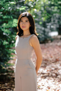 A woman with medium-length dark hair stands outdoors on a leaf-covered path, wearing a sleeveless beige dress. She faces the camera with a slight smile, as if ready for a Rachel Heng interview, surrounded by green trees in sunlight.
