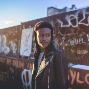 A young person in a black leather jacket and hoodie stands outdoors in front of a graffiti-covered wall, looking to the side with a neutral expression—as if awaiting a tyriek white interview. The sunlight creates a soft glow in the background.