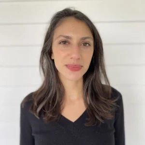 A woman with long brown hair and a black V-neck sweater stands in front of a white paneled wall, looking straight at the camera with a neutral expression, reminiscent of an Emma Ramadan interview portrait.
