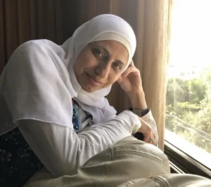 A woman wearing a white hijab and long-sleeved top rests her head on her hand while sitting by a sunlit window, looking at the camera with a gentle smile, reminiscent of moments from the Dareen Tatour interview.