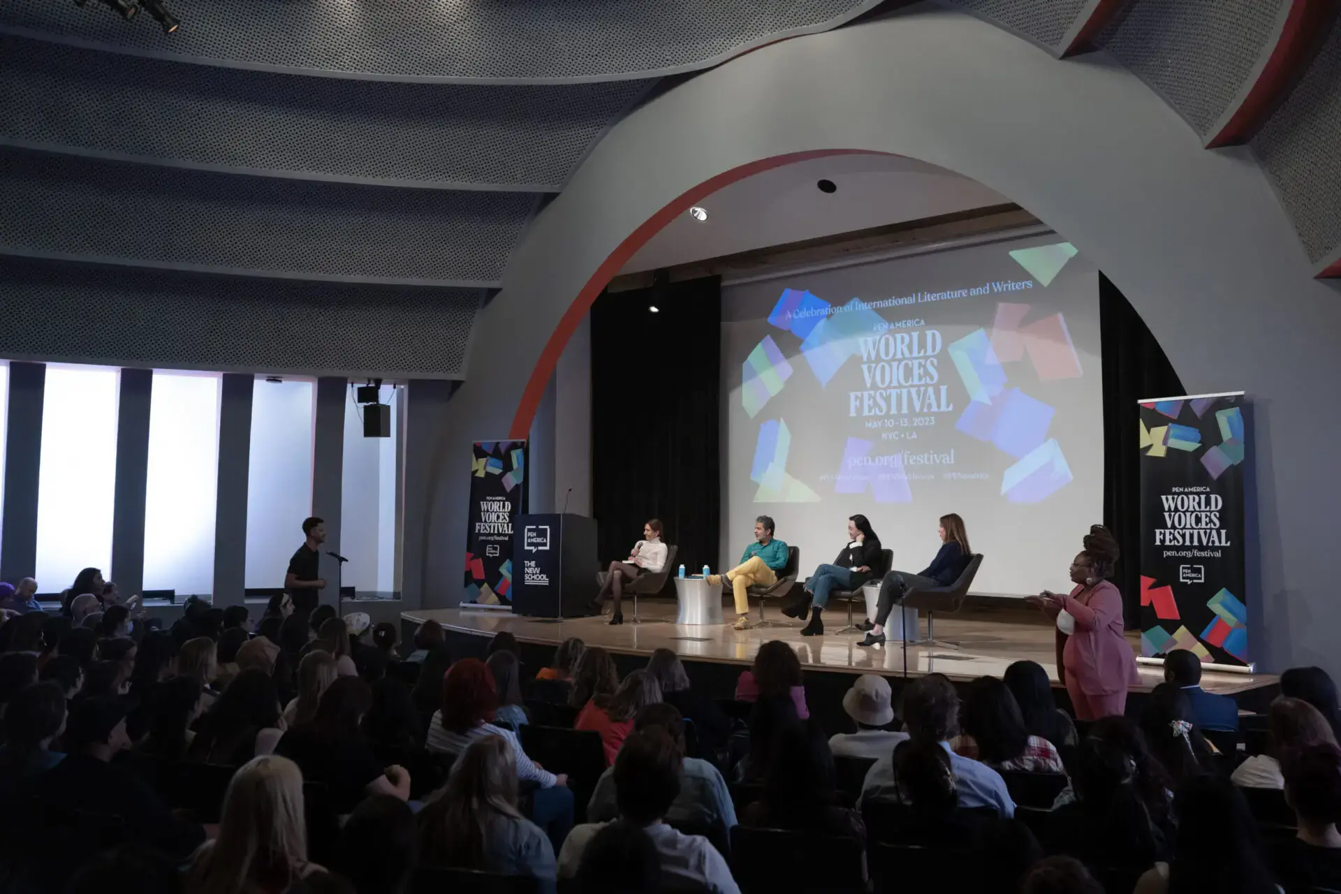 A panel of four speakers sits on stage under a large arch, addressing an audience at the World Voices Festival 2023. A screen behind them displays the event name, while the room is filled with engaged attendees.