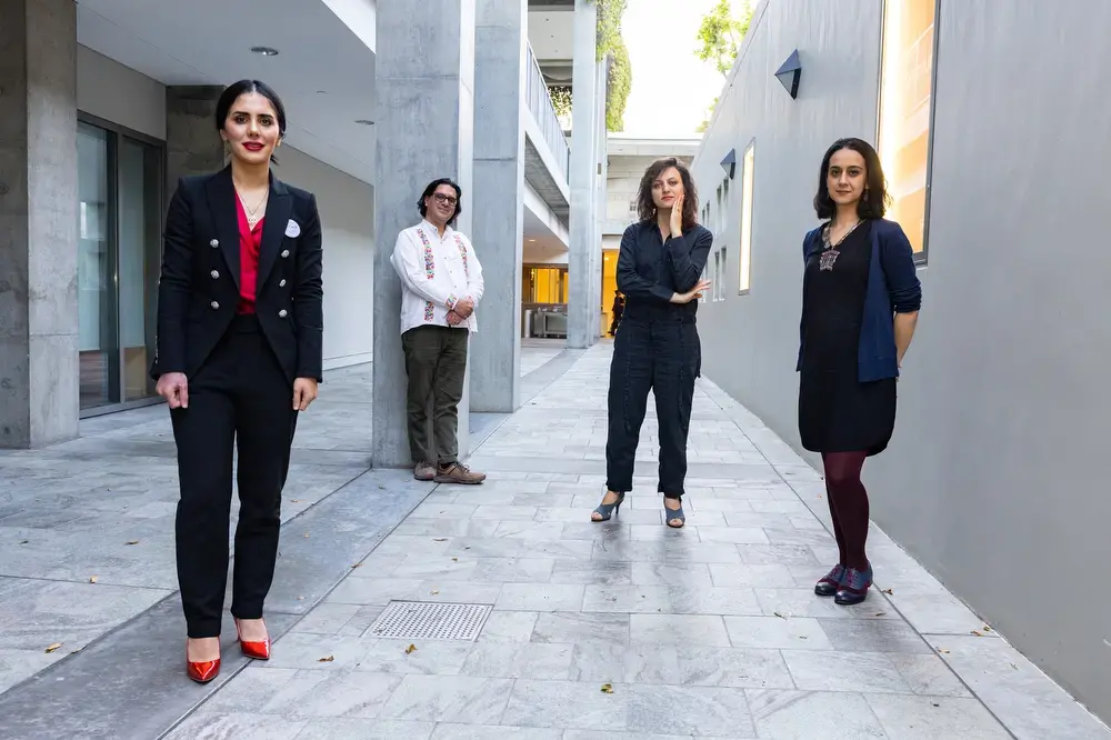 Four women stand spaced apart in a modern outdoor corridor with gray walls and concrete floors. Three are closer to the camera, while one stands farther back by a column—all confidently facing forward at the World Voices Festival 2023.