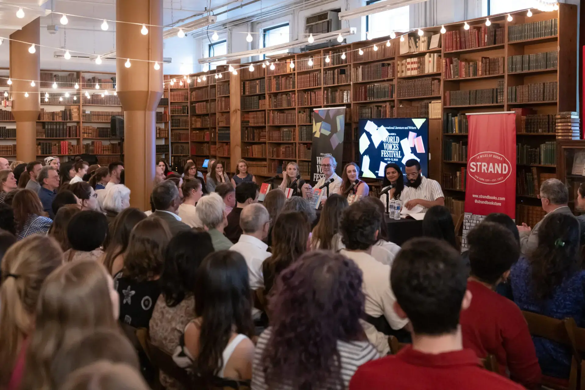 A large audience listens to a panel of speakers at a book event in a library, with shelves of books, string lights above, and banners for the Strand bookstore and World Voices Festival 2023 visible.