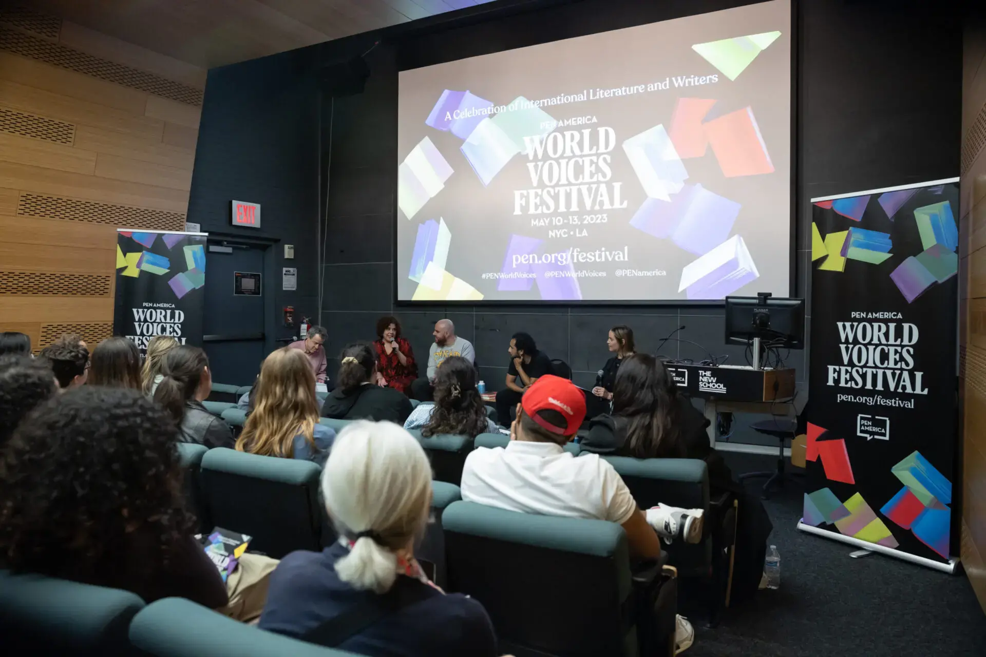 A diverse panel of speakers sits on stage in front of an audience, with a large screen behind them displaying World Voices Festival 2023 and event details.