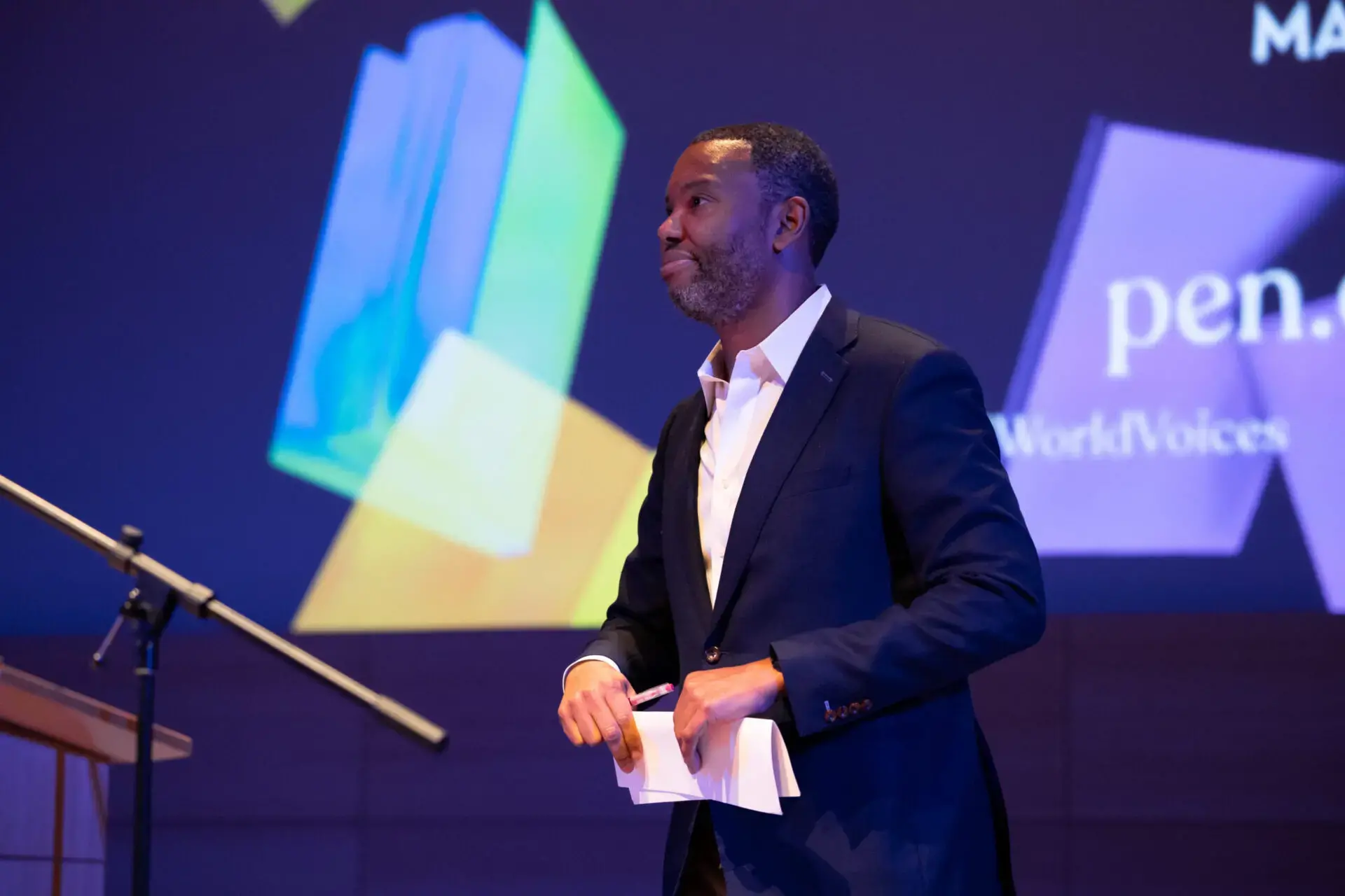 A man in a dark suit holds papers and stands on stage in front of a microphone, with colorful geometric shapes and the words pen., WorkVoices, and World Voices Festival 2023 displayed on a screen behind him.