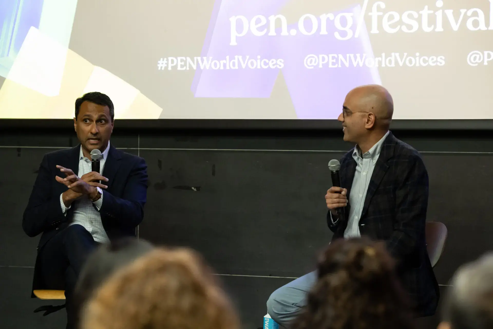Two men sit on a stage in conversation at the World Voices Festival 2023; one gestures while speaking, the other listens with a microphone. The background displays pen.org/festival and #PENWorldVoices on a screen.