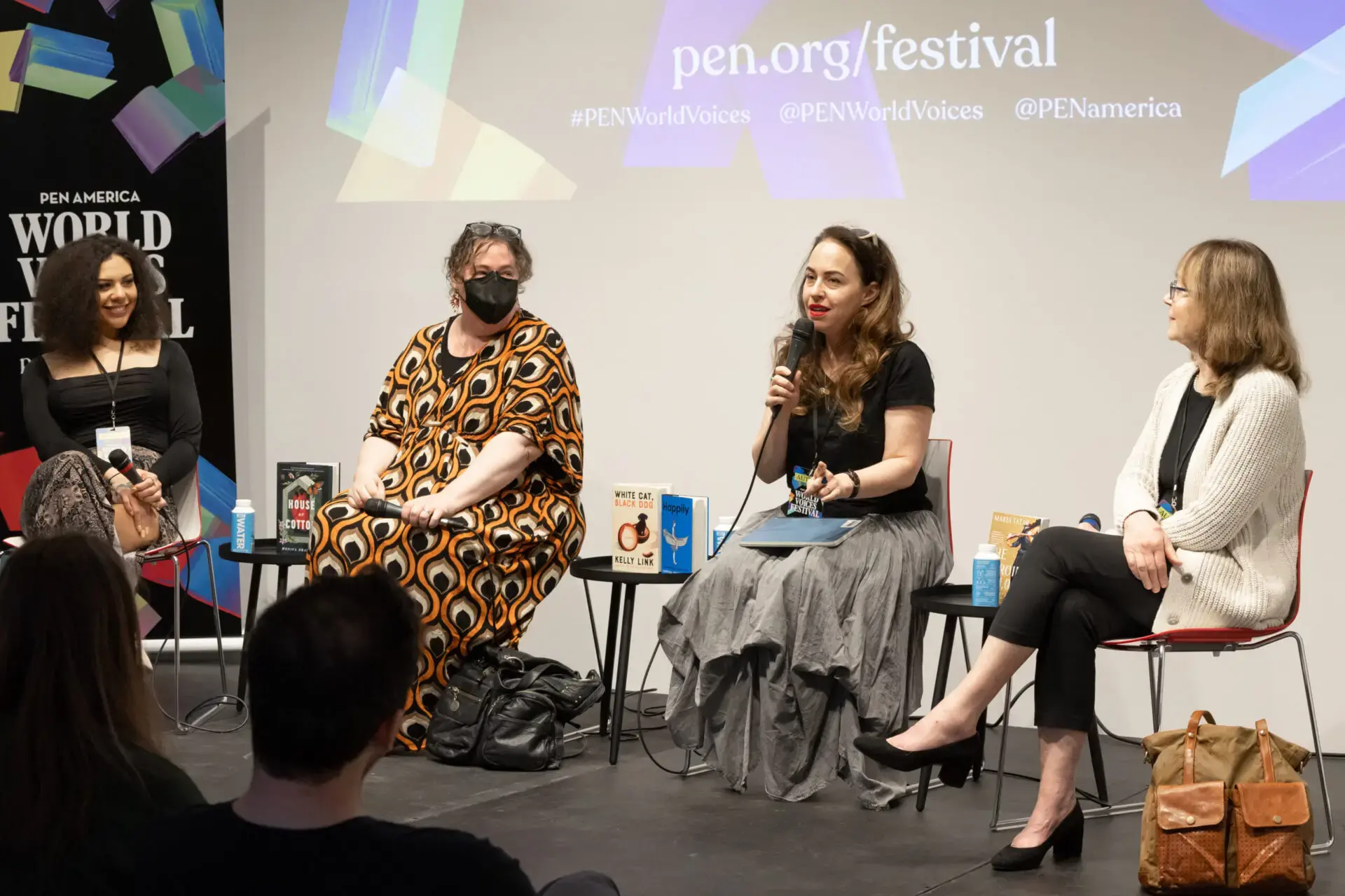 Four women sit on stage at a literary panel during the World Voices Festival 2023. One speaks into a microphone as others listen. Books are displayed on the table between them, and a festival banner is projected behind them.