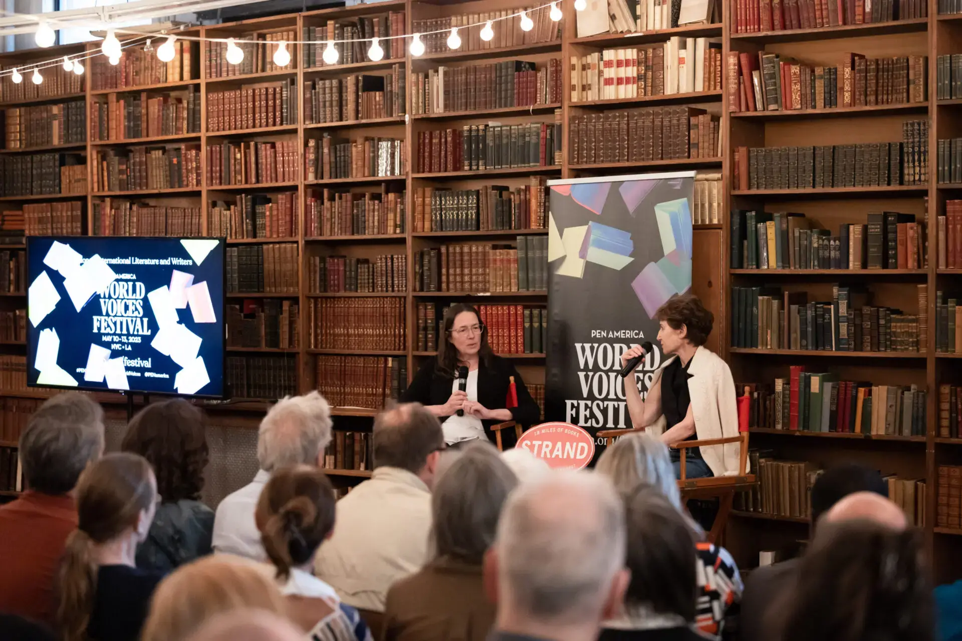 Two women speak onstage in front of bookshelves at a World Voices Festival 2023 event, as an audience watches. A large banner and a screen with the festivals 2023 logo are visible beside them.