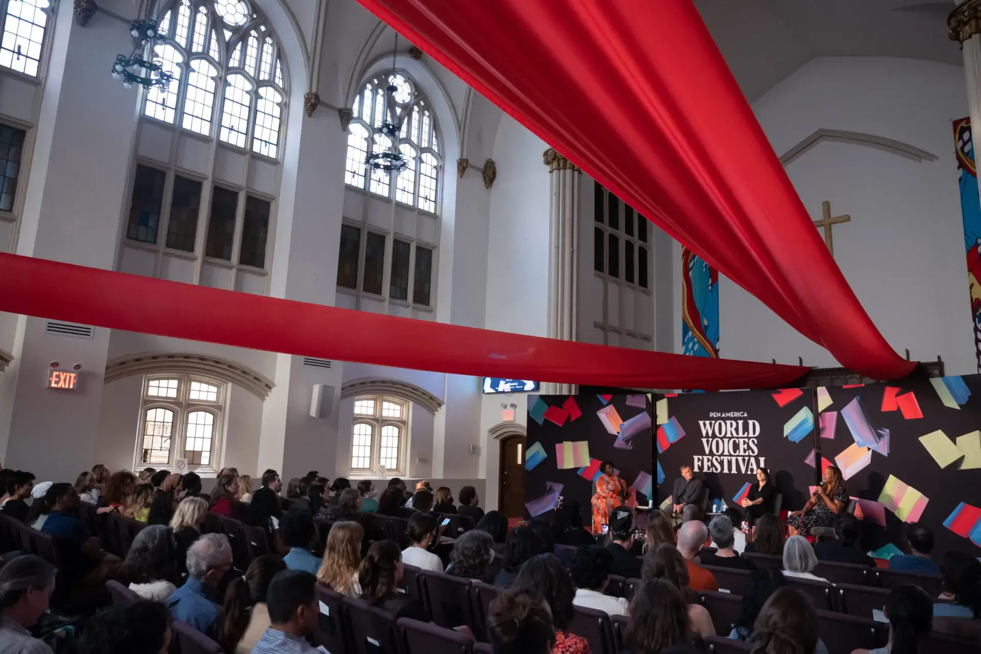 A diverse audience sits in a church-like venue with tall arched windows, watching speakers on stage at the World Voices Festival 2023. Colorful decorations and a large red ribbon hang from the ceiling.