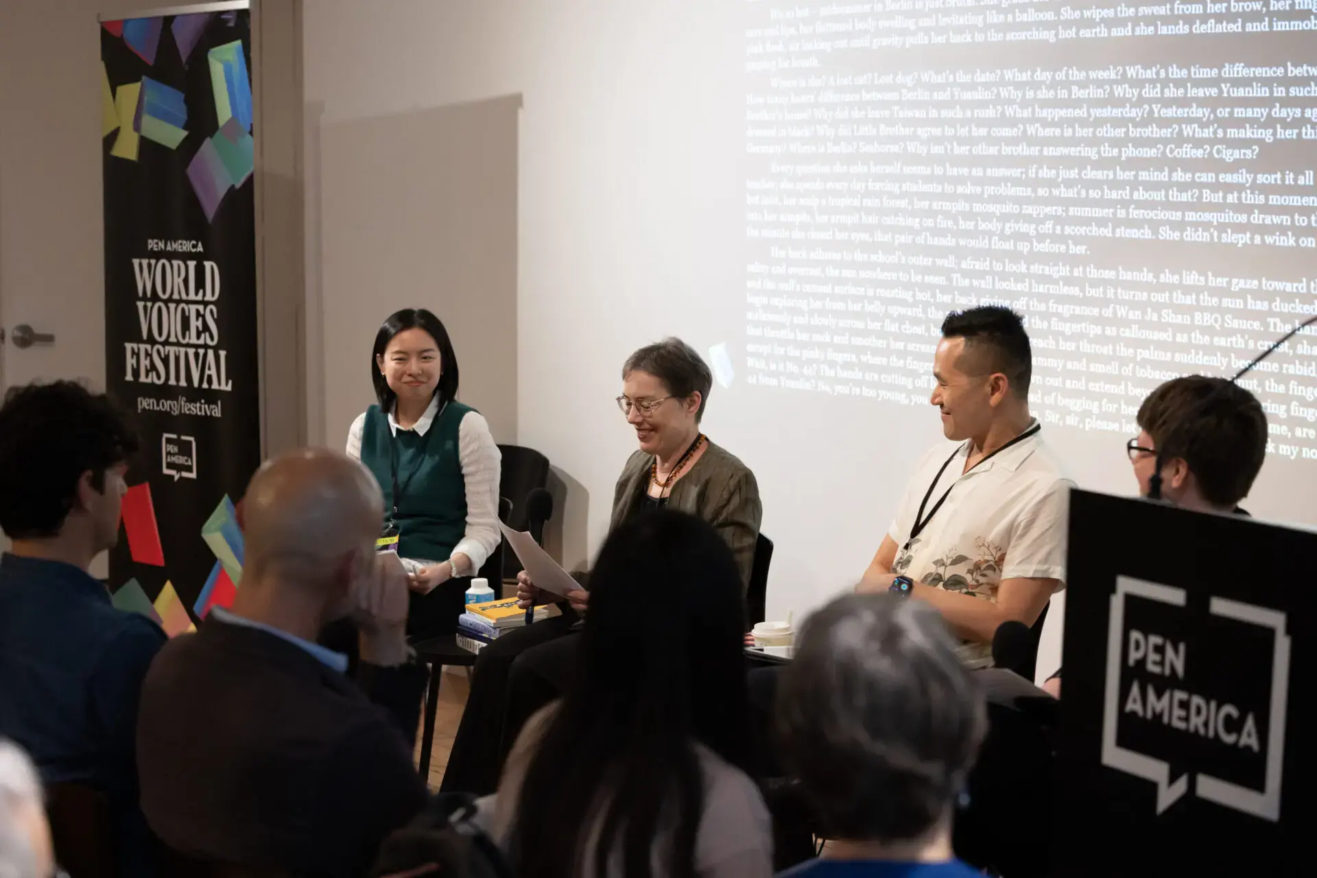 Four panelists sit before an audience at the World Voices Festival 2023. A woman in the center reads from a paper while text is projected behind them. Audience members listen attentively throughout the engaging event.