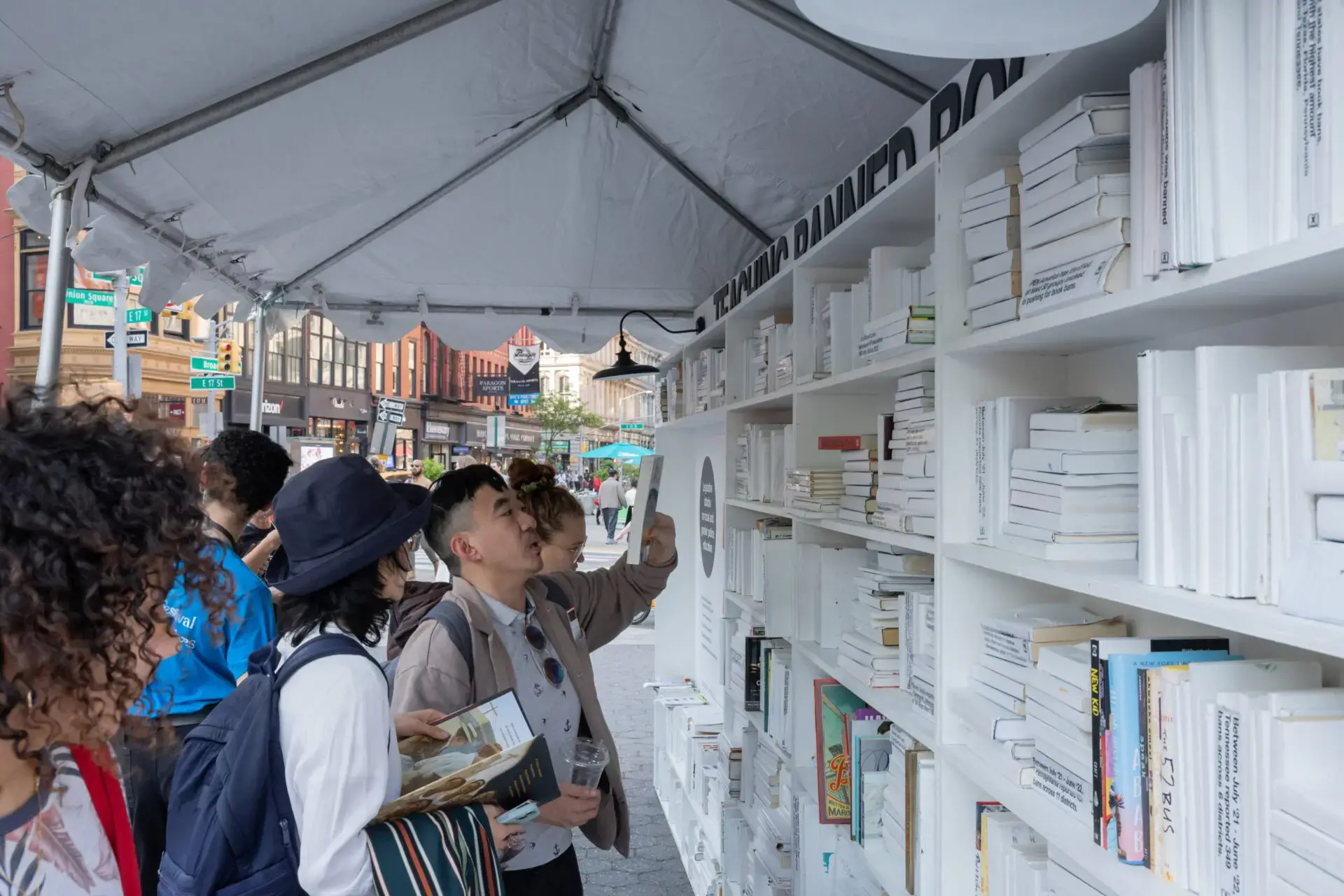 People browse and select books from white shelves under a canopy at the World Voices Festival 2023 outdoor book fair, with city buildings and street signs visible in the background.
