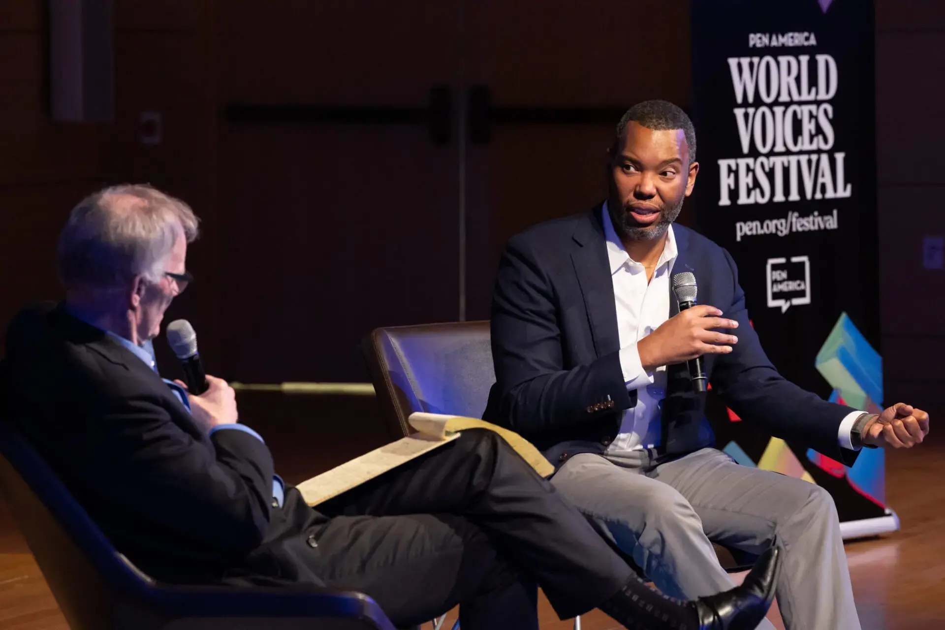 Two men sit on stage in armchairs, engaged in conversation with microphones. One holds a notepad. A banner in the background reads PEN America World Voices Festival 2023, highlighting this public event or talk.