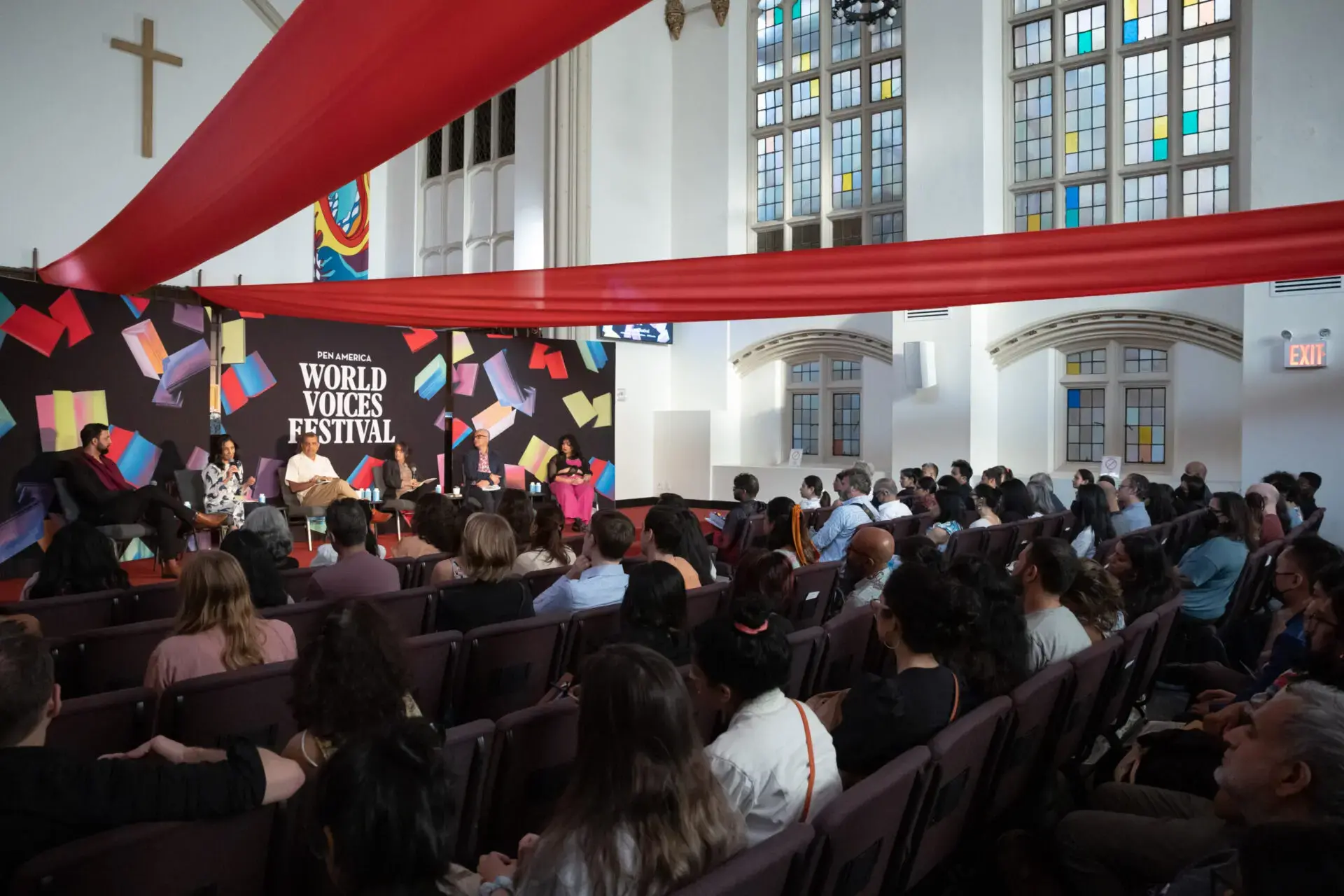 A diverse audience sits in rows inside a church-like venue, watching a panel discussion on stage at the World Voices Festival 2023. Colorful banners and large windows decorate the lively space.
