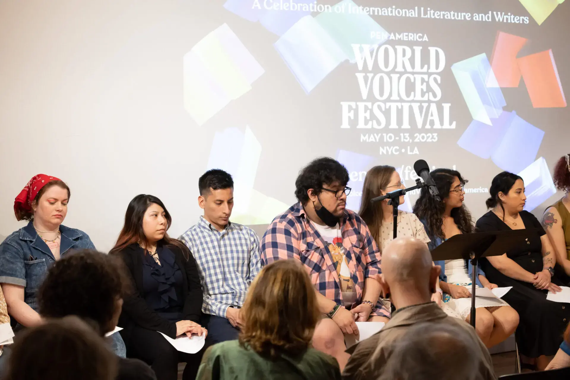 A group sits on stage before a screen announcing World Voices Festival 2023, May 10-13, NYC + LA. Some listen intently while others look ahead; audience members can be seen in the foreground.