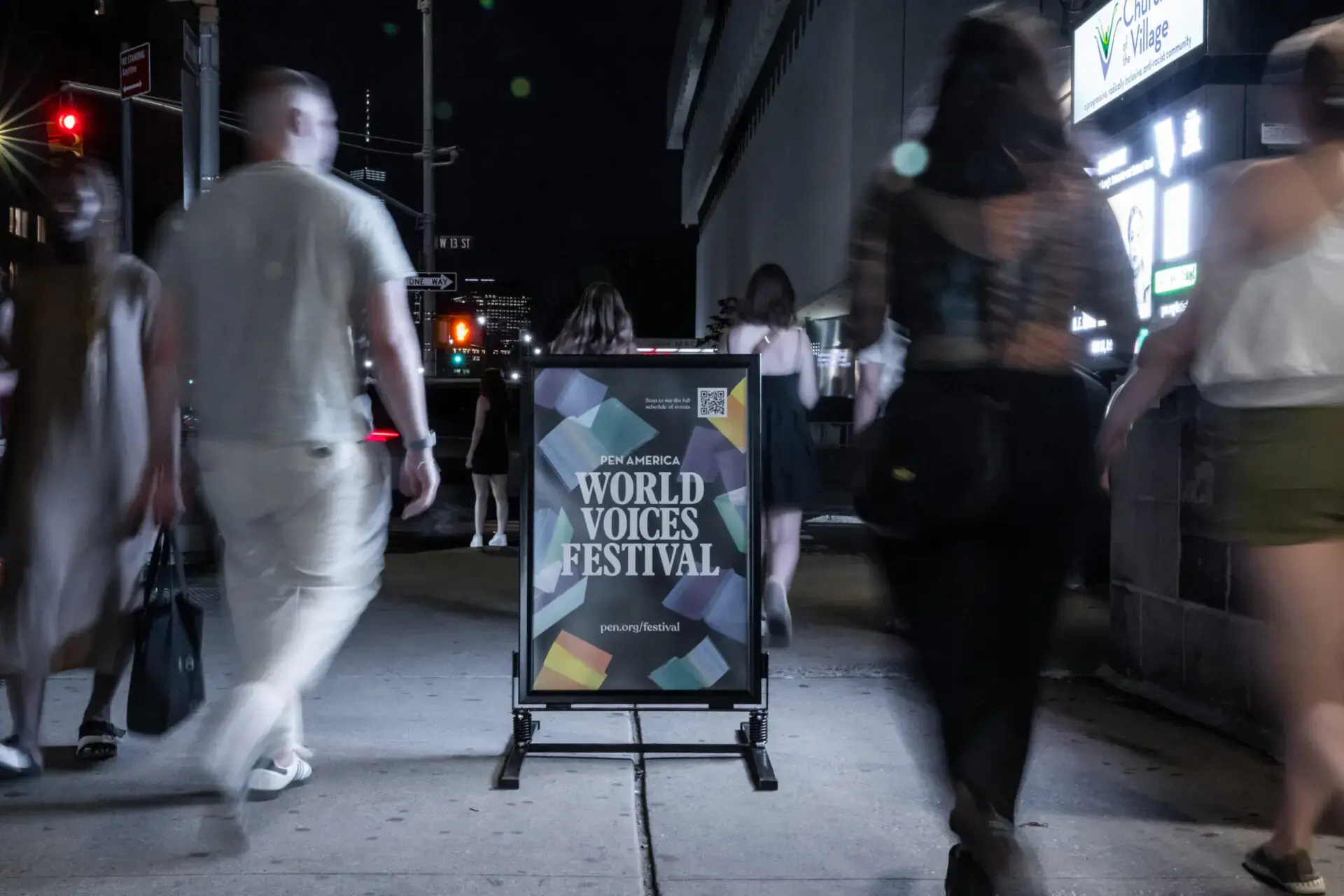 People walk on a city sidewalk at night, passing a sign that reads “PEN America World Voices Festival 2023.” The scene is dimly lit, with blurred figures and urban streetlights in the background.