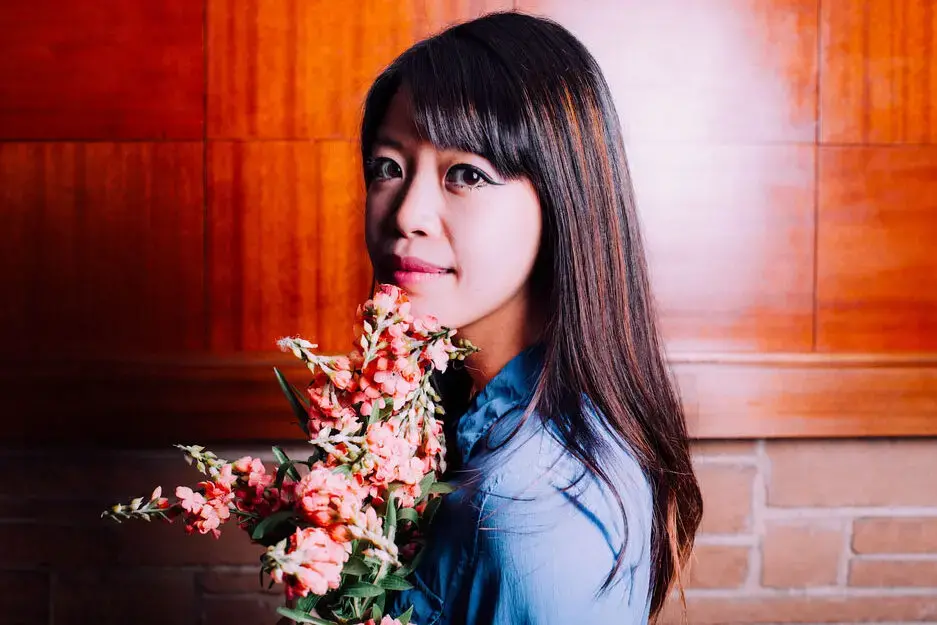 A woman with long dark hair and a blue shirt holds a bouquet of pink flowers, standing in front of a wooden and brick wall, looking at the camera with a gentle expression—reminiscent of the calm tone in the Jane Wong interview.