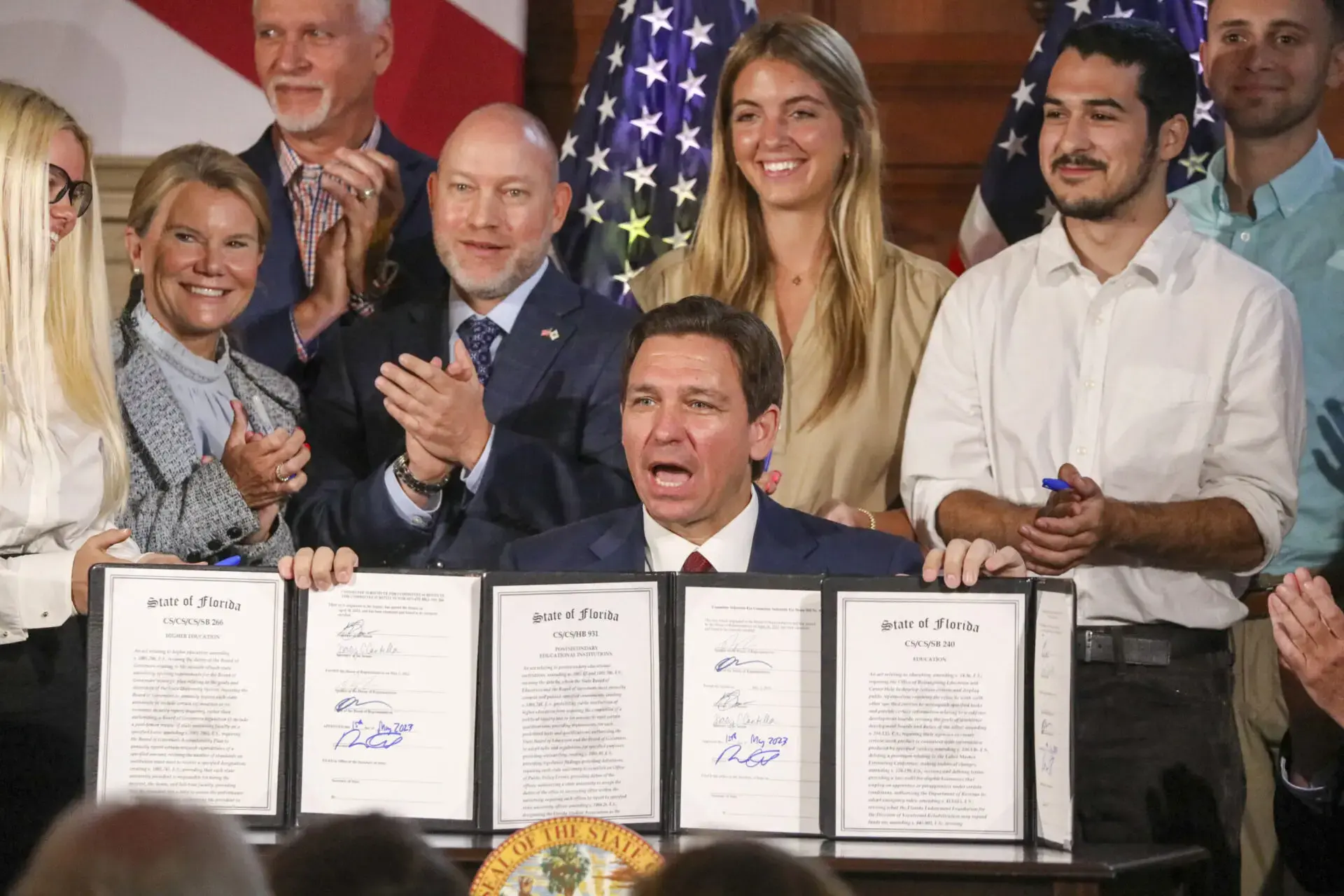 A man in a suit sits at a desk, holding up four signed official documents, while smiling people stand behind him. American flags are visible in the background.