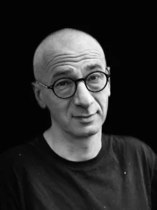 A person with a shaved head and round glasses wearing a black shirt looks at the camera with a slight, inquisitive expression against a plain dark background.