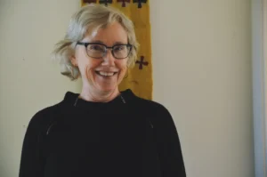 A smiling older woman with short gray hair and glasses stands indoors against a light-colored wall with a yellow fabric featuring red and black cross patterns hanging behind her.