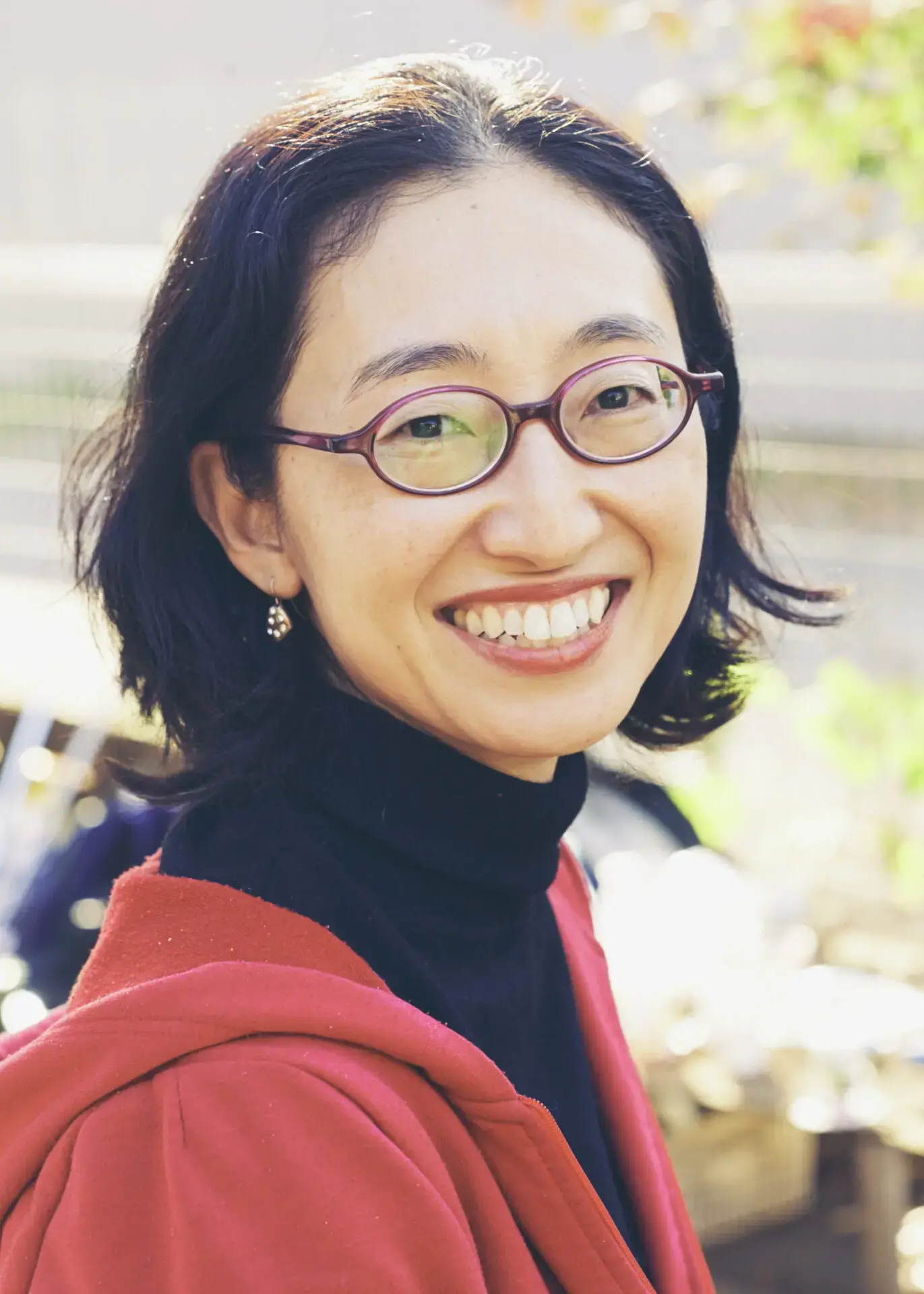A woman with chin-length black hair, red-framed glasses, and a black turtleneck smiles brightly outdoors. She is wearing a red jacket and sunlight softly illuminates her face.