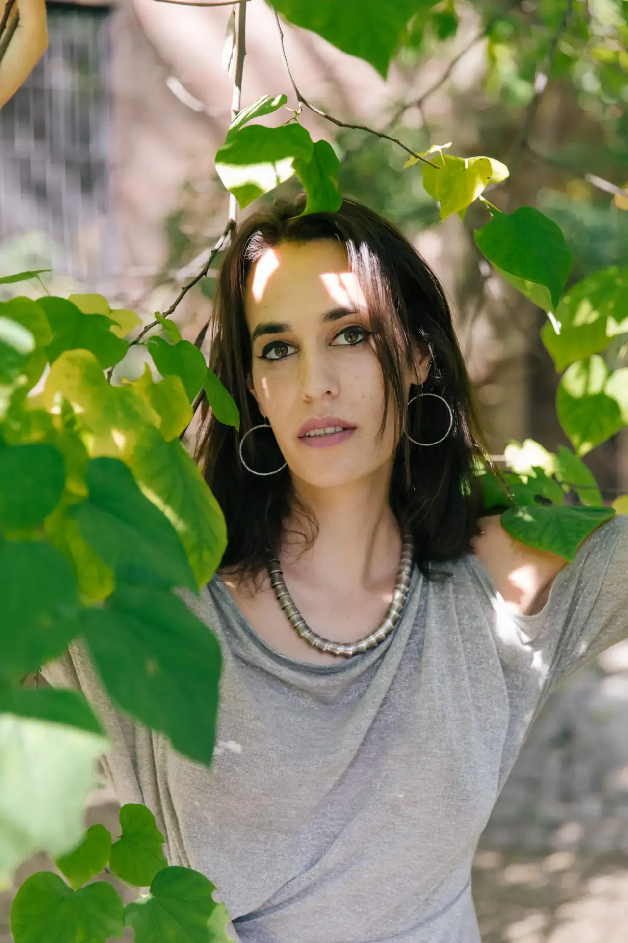 A woman with dark hair and hoop earrings stands outdoors among green leaves, wearing a gray top and a beaded necklace. Sunlight filters through the leaves, casting shadows on her face.