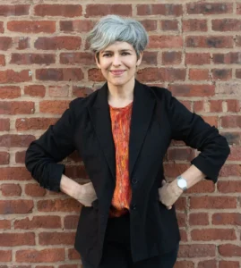 A person with short gray hair wearing a black blazer, orange shirt, and watch stands smiling with hands on hips in front of a brick wall.