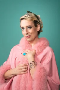 A person with short blond hair, wearing a pink fur-trimmed shawl, a large blue ring, and a silver bracelet, stands against a teal background and smiles softly at the camera—evoking the poise of a Grace Lavery interview.