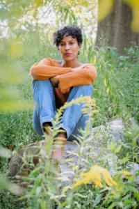 A person with short curly hair sits barefoot on a rock in a green, grassy area, wearing a bright orange sweater and blue jeans, as if deep in thought after an Aracelis Girmay interview, arms wrapped around their knees and surrounded by foliage.