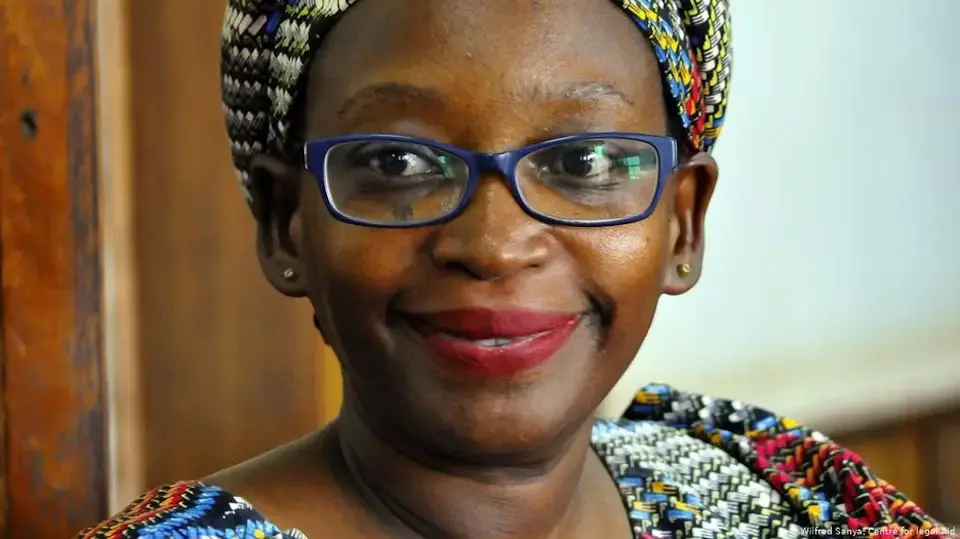 A woman wearing glasses, a colorful patterned headwrap, and matching top smiles softly, looking slightly to the side—her expression reminiscent of the warmth seen in a Stella Nyanzi interview. The softly blurred background highlights her cheerful face.