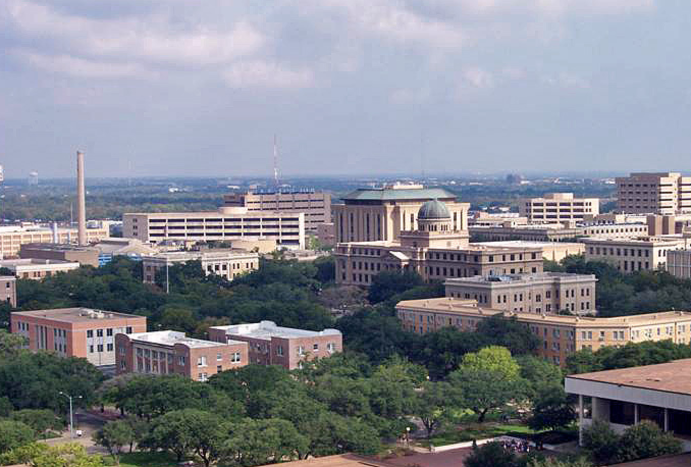 A wide view of a university campus with several large, historic buildings surrounded by trees, set against a backdrop of more modern structures under a partly cloudy sky.