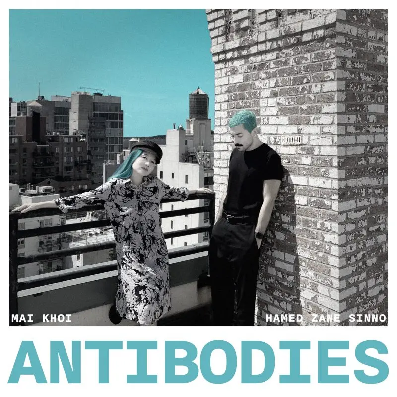 Two people with blue hair stand on a city rooftop—Mai Khoi leans against a railing in a floral dress and hat, while Hamed Sinno stands by a brick wall in black. The text ANTIBODIES appears at the bottom, their names above.
