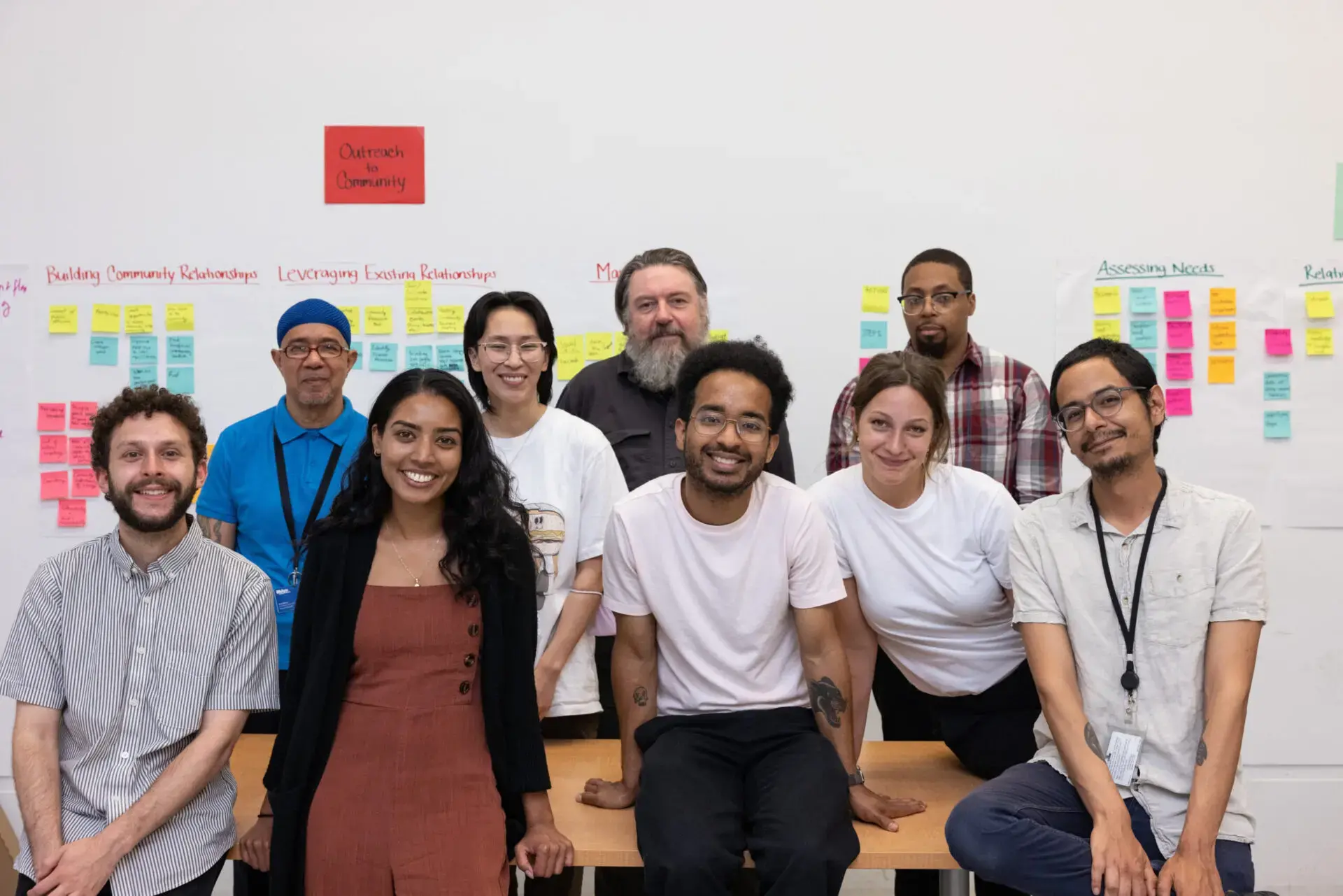 A diverse group of nine people, including Valentina Flores, pose together and smile in a brightly lit room, standing and sitting in front of a wall covered with colorful sticky notes and Justice Initiatives project planning posters.