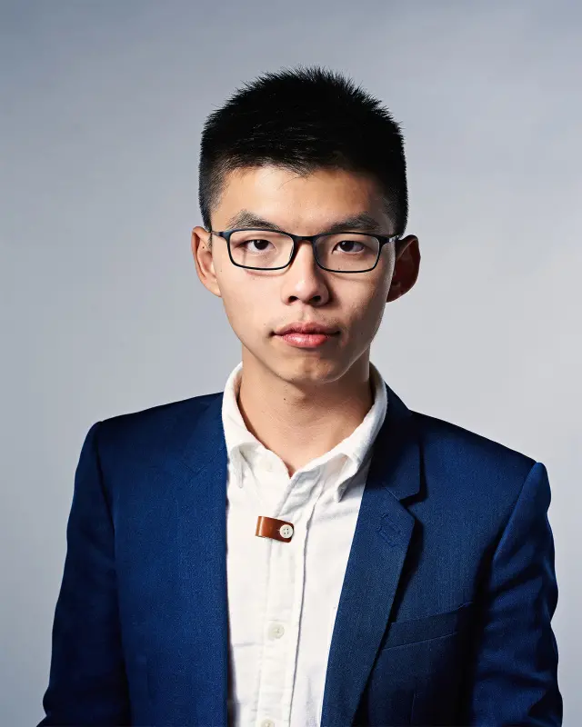 A young man wearing glasses, a white shirt, and a blue blazer stands against a plain, light-colored background, looking directly at the camera with a neutral expression.