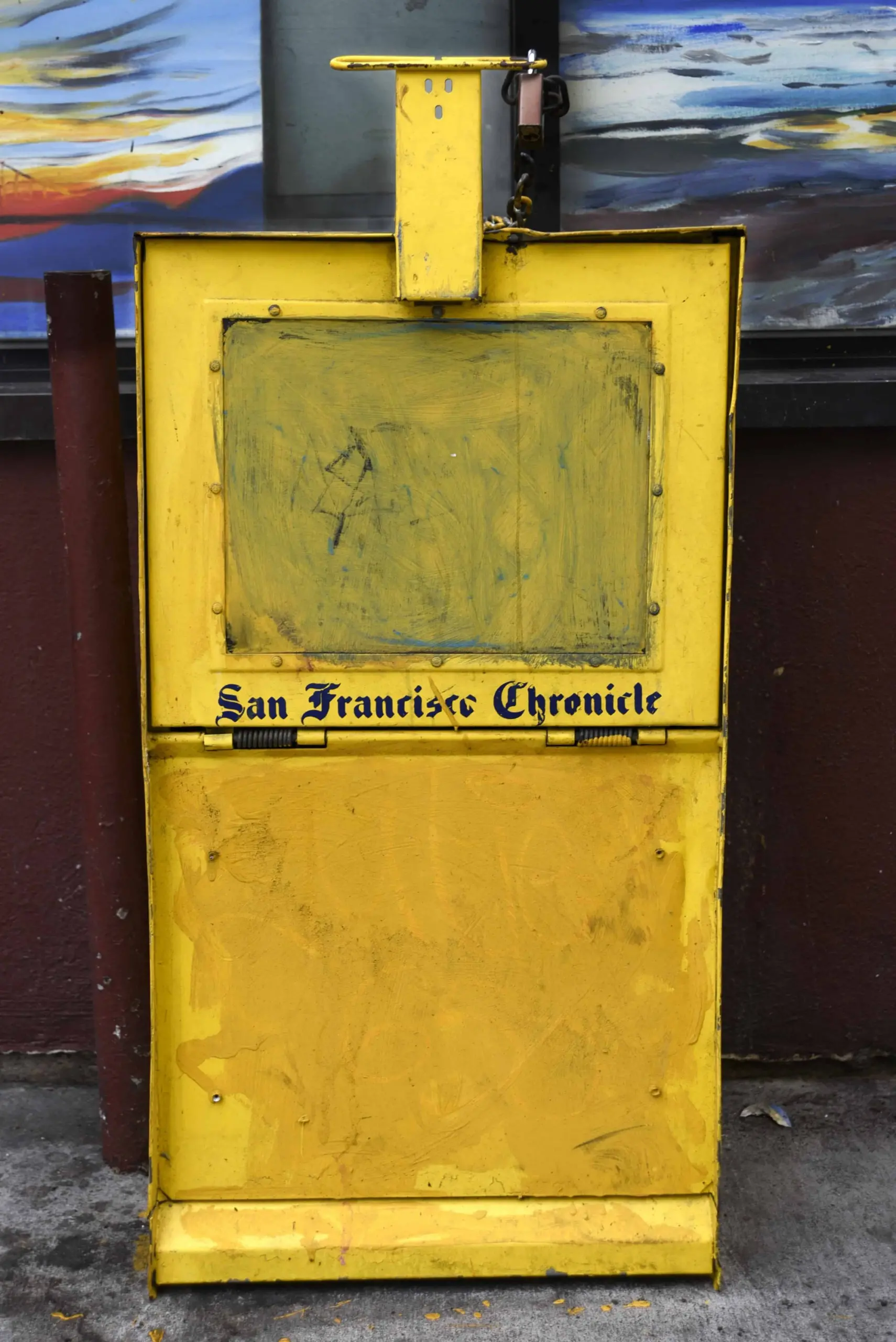 A weathered yellow newspaper vending box for the San Francisco Chronicle stands on a sidewalk in front of a burgundy wall, with grime and faded paint visible on its surface.