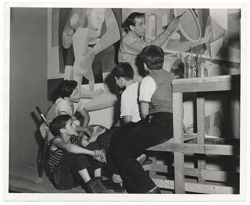 A man gestures toward a large abstract mural while four boys sit and listen attentively, surrounded by art supplies in a studio setting.