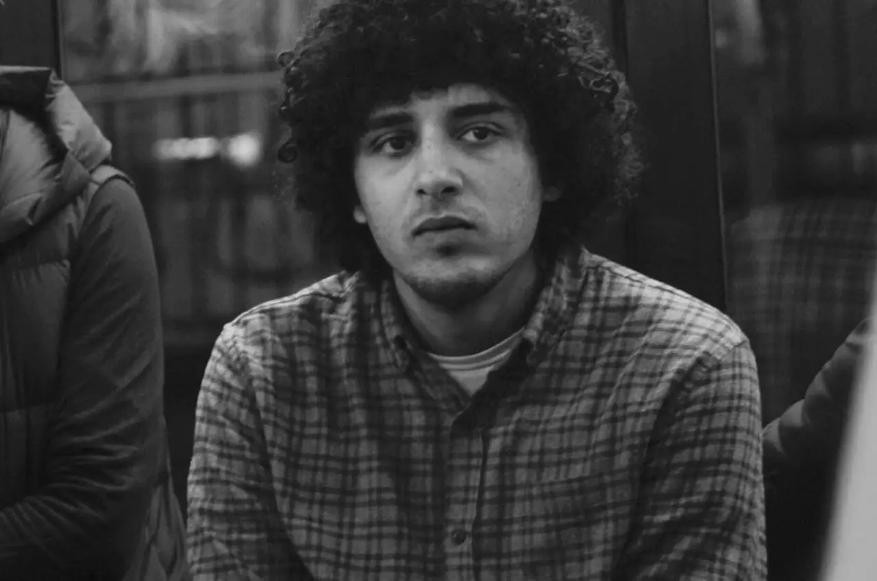 Black and white photo of a young man with curly hair wearing a plaid shirt, looking directly at the camera with a neutral expression. A blurred figure and background are visible behind him.