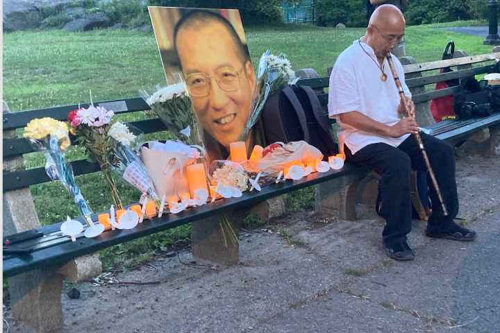 A man plays a flute on a park bench beside flowers, candles, and a large portrait of an older man with glasses, set up as part of a memorial display.