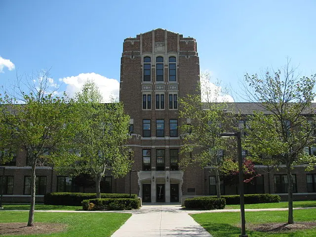 A tall, brick academic building with large windows and an arched entrance stands behind a path, flanked by green trees and bushes, under a blue sky with scattered clouds.