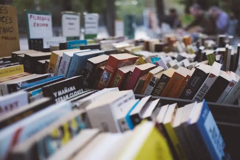 Rows of books displayed on tables at an outdoor book market, with various colorful covers and signs in the background indicating different genres and sections.