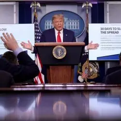 A man stands at a podium with the presidential seal, speaking at a press briefing. People in the audience raise their hands. Two screens behind him display slides titled 30 Days to Slow the Spread.