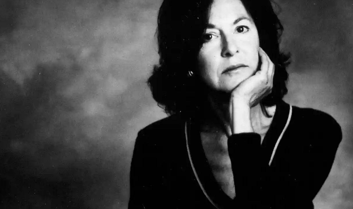 Black and white photo of a woman with dark, shoulder-length hair, resting her chin on her hand, gazing thoughtfully at the camera, wearing a dark top with a light-colored trim. The background is plain and softly blurred.