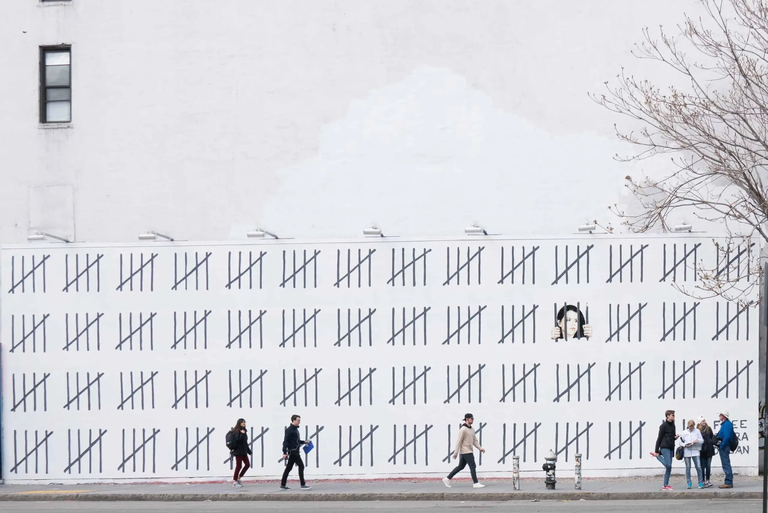 Six people walk past a large white wall covered in tally marks; two people appear to be looking out of a window on the wall, with a mostly bare tree branch extending into the frame from the right.
