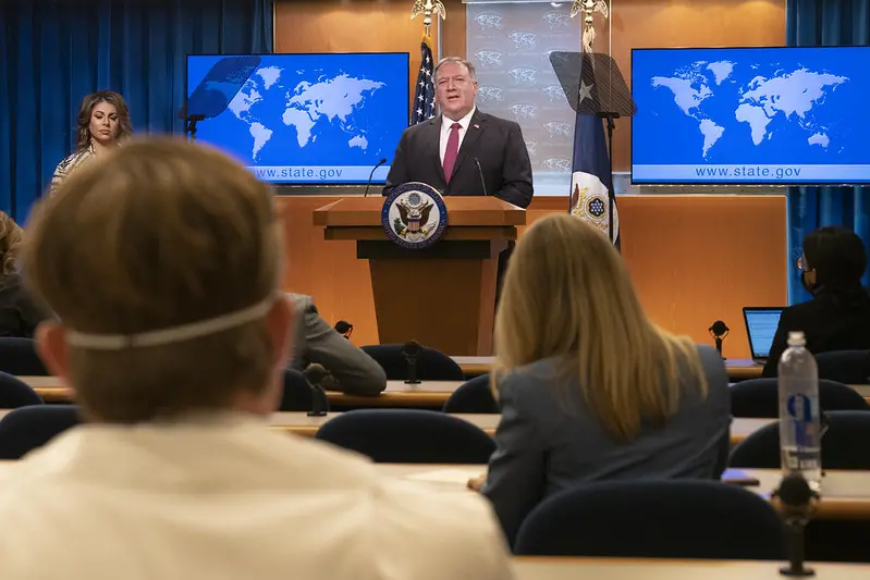 A man stands at a podium with the U.S. State Department seal, speaking at a press conference. People sit facing him, and two screens behind him display a world map and “www.state.gov.”.