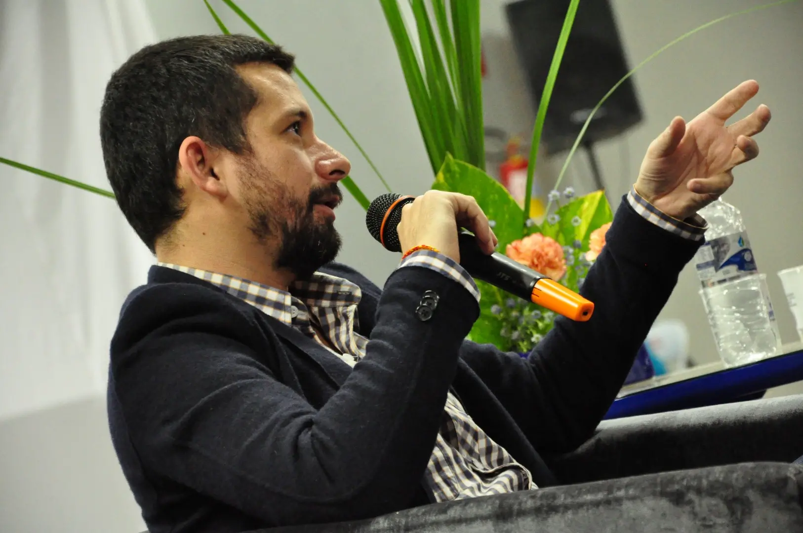 A man with short dark hair and a beard speaks into a microphone while sitting on a gray chair, gesturing with his hand. He wears a checkered shirt and dark blazer. There are flowers and a water bottle in the background.