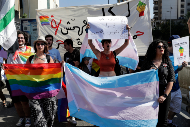 A group of people at a protest hold rainbow and transgender pride flags. Some carry signs with Arabic writing, and a large banner is displayed in the background. Buildings are visible behind them.