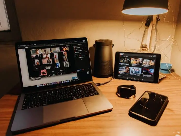 A laptop showing a video call, a tablet displaying YouTube thumbnails, a smartwatch, a smartphone, a speaker, and a desk lamp are arranged on a wooden desk in a cozy, warmly lit room.